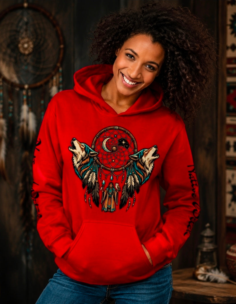 A smiling woman with curly dark hair wears a bright red pullover hoodie featuring a large dreamcatcher and wolf graphic on the front, paired with blue jeans, set against a rustic dark wood background with dreamcatchers.
