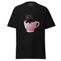 A black short-sleeve t-shirt featuring a graphic of a black cat sitting inside a pink teacup with the words 'MAGIC BREW' printed on the front.